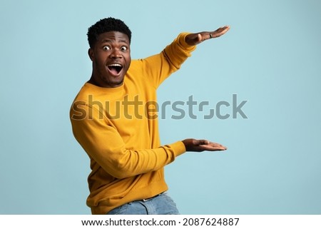 Amazed african american young guy in yellow shirt and jeans holding something invisible in his hands and showing astonishment, blue studio background, panorama with copy space