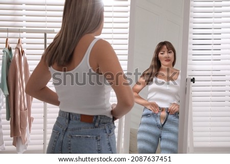 Young woman trying to put on tight jeans near mirror at home Royalty-Free Stock Photo #2087637451