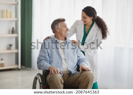 Portrait of happy senior man in wheelchair and his female nurse at retirement home. Mature male patient with disability and his caregiver posing and smiling, looking at each other indoors Royalty-Free Stock Photo #2087641363