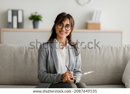 Professional therapist concept. Portrait of young female arab psychologist sitting on couch, holding clipboard with pen and writing, posing and smiling at camera