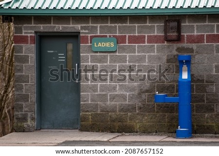 The entrance to a ladies washroom in a park. The lavatory has a green door, brick wall, wooden sign, and metal roof. There's a blue tri-level outdoor pedestal tubular pet water refilling station.