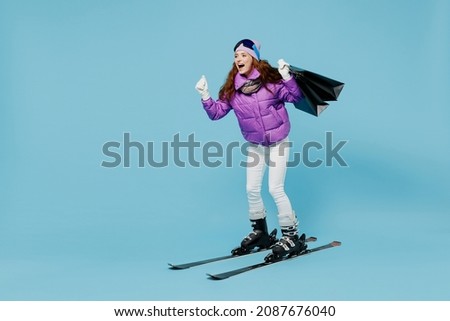 Full body skier woman in warm purple padded windbreaker jacket ski goggles mask spend weekend in mountains holding package bags with purchases after shopping isolated on plain blue background studio.
