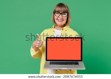 Elderly woman 50s in glasses yellow shirt hold use work on laptop pc computer with blank screen workspace area show thumb up isolated on plain green background studio portrait People lifestyle concept