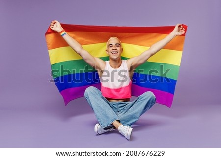 Full body young latin gay man with make up wearing bright pink top sitting hold rainbow striped flag isolated on plain pastel purple background studio portrait. People lifestyle fashion lgbtq concept Royalty-Free Stock Photo #2087676229