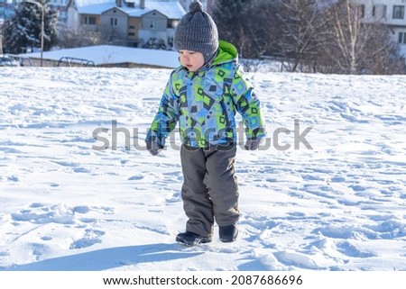 A child with a serious expression on his face in winter clothes jackets, pants, hat and boots in winter on the white snow on the street and in the park in nature plays winter fun. Royalty-Free Stock Photo #2087686696