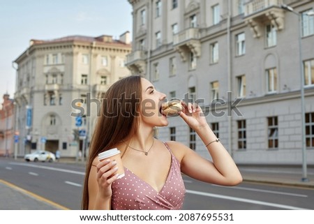 ttractive girl with long hair takes a bite of a cake and holds a paper cup of coffee. photo shoot in the city. Royalty-Free Stock Photo #2087695531