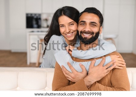 Happy lovely young Indian couple together at home, ethnic young wife hugging from behind her husband, sitting and resting on sofa in modern apartment, portrait of romantic multiracial couple in love Royalty-Free Stock Photo #2087699998