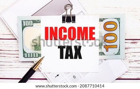 On a light wooden table, there is a white calculator, a pen, bills, and a sheet of paper under a black paper clip with the text INCOME TAX. Business concept