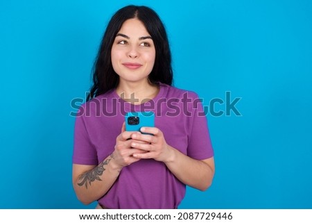 Happy Caucasian woman wearing purple T-shirt over blue background listening to music with earphones using mobile phone.