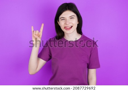 Caucasian woman wearing purple T-shirt isolated over studio background doing a rock gesture and smiling to the camera. Ready to go to her favorite band concert.