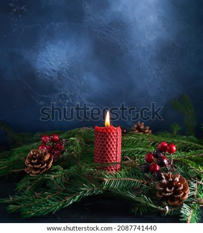 A red candle made of natural wax burns surrounded by green fir branches. Cones and red berries in the background. Dark blue textured background. New Year's card