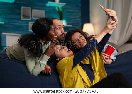 Group of friends smiling at camera while happy woman taking selfie sharing picture on social media, having fun celebrating wekeend at home. Multi-ethnic people laughing making funny expression