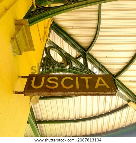 A brown sign with the Italian word for exit written in yellow letters is attached to a yellow wall under the roof of the Bagni di Lucca train station with green girders.