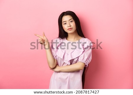 Young smug asian girl looking cool and pointing finger left at logo, advertising product on pink romantic background Royalty-Free Stock Photo #2087817427