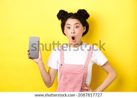 Online shopping concept. Excited stylish asian girl say wow, showing empty cell phone screen and look amazed, yellow background