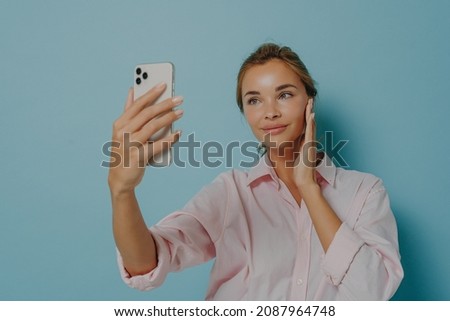 Beautiful woman with tender expression looks at smartphone camera takes selfie of herself wears formal shirt isolated over blue background talks on video app. Communication and online technology