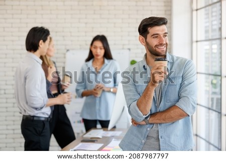 Positive male worker with takeaway hot drink standing near group of multiethnic coworkers discussing business project in light conference room