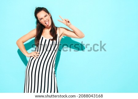 Young beautiful smiling female in trendy summer zebra dress. carefree woman posing near blue wall in studio. Positive brunette model having fun and shows peace sign. Cheerful and happy