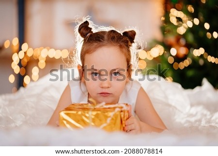 a kid girl in a white dress sits on the floor on a rug with a gift in gold wrapping paper. tradition to give gifts to children for the New Year and birthday. Santa Claus. children's toy store.
