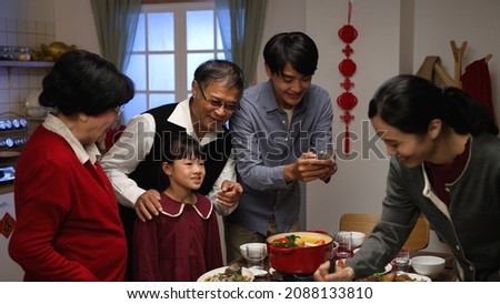 excited asian male father taking photos of family getting together for chinese new year dinner by dining table with mobile phone while young mother is cooking hot pot