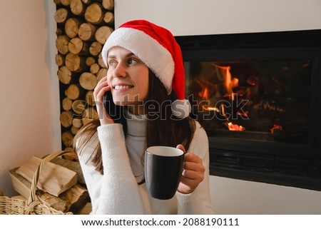 Smiling woman in white sweater and Christmas hat sitting on floor near fireplace at home talking on mobile phone, girl holding hot cup coffee or tea. Girl calling to service for making delivery online