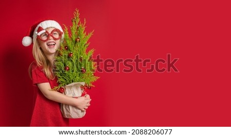a little girl in a red dress, a Christmas cap and carnival glasses holds a green live Christmas tree with New Year's decorations in her hands. The child laughs happily, shows his teeth. Copy space