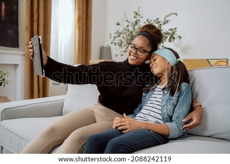 Mom sits on couch with daughter and embraces her, girls smile, woman pulls out tablet far in hand to take commemorative selfie of them on social media