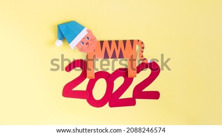 A bright tiger made of paper in a Christmas hat on a light background. Minimalistic Christmas card paper numbers 2022 and origami tiger on a colored background