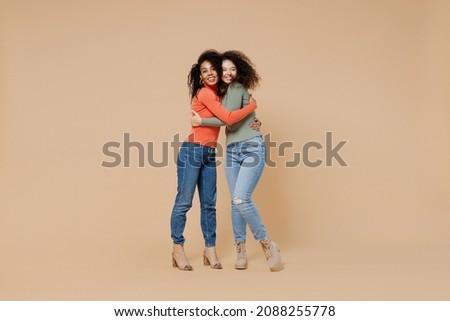 Full size body length two friendly fun young curly black women friends 20s wear casual shirts clothes meeting greeting hugging looking camera isolated on plain pastel beige background studio portrait