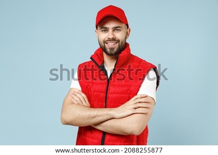 Professional calm delivery guy employee man in red cap white T-shirt vest uniform workwear work as dealer courier looks camera isolated on pastel blue color background studio portrait. Service concept