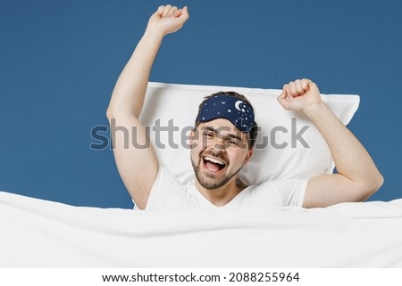 Young happy man 20s wear pajamas jam sleep mask rest relaxing home with lies wrap covered under blanket duvet with look camera yawning isolated on dark blue background Good mood night bedtime concept Royalty-Free Stock Photo #2088255964