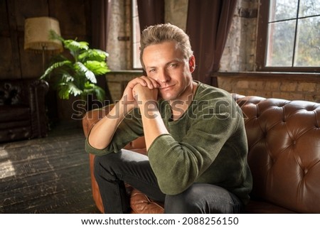 Attractive Male of 35s with Loving Eyes in Casual Clothes Sits on a Soft Leather Sofa in his Luxury Living Room of his Country House. Close-up. . High quality photo Royalty-Free Stock Photo #2088256150