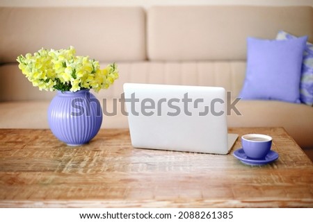 Open laptop, cup of coffee and very peri vase with beautiful yellow flowers on retro wood textured table in minimalistic living room with beige couch in background. Freelance workplace at home Royalty-Free Stock Photo #2088261385