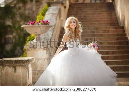 A naughty blonde bride, with cool make-up in a wedding dress. Stands against the background of the old church and smiles. Bride beautiful wedding flowers bouquet, gorgeous woman. Husband and wife. Royalty-Free Stock Photo #2088277870