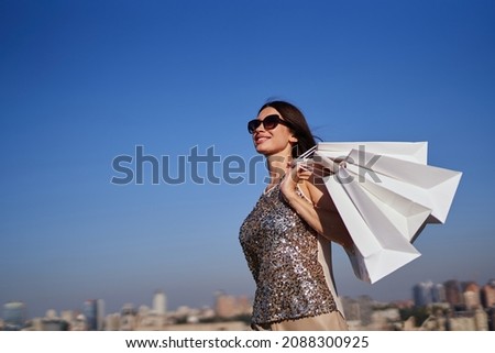 Attractive caucasian fashionable woman with white shopping bags going in front of blue sky and urban city background. Glam shopaholic brunette girl in sunglasses. Retail, consumerism or sale concept Royalty-Free Stock Photo #2088300925