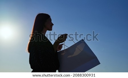 Silhouette of happy fashionable long hair woman looking at shopping bags in sunset, image against sun and blue sky. Consumerism, shopping, sale or black friday concept. High quality photo