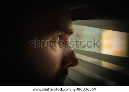 A man in a dark room looks out the window through the blinds. Covert surveillance. Close-up Royalty-Free Stock Photo #2088336859