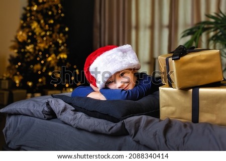 Portrait of a beautiful boy in a Santa hat on a bed with a gift on the background of a Christmas tree with bokeh lights.