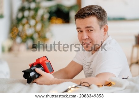 man in white t-shirt with camera in red silicone case against background of Christmas decorations. work of professional photographer during Christmas holidays. accessories for photographic equipment.