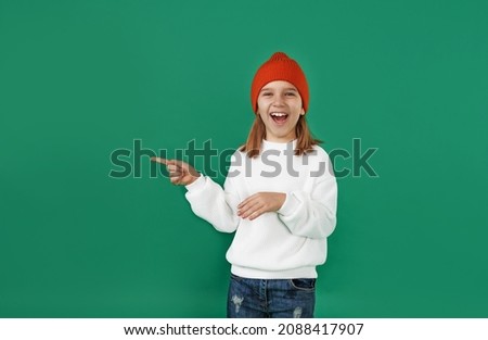 a little girl in a white sweater and a red hat laughs and points her finger to the side on a green isolated background. children's emotions