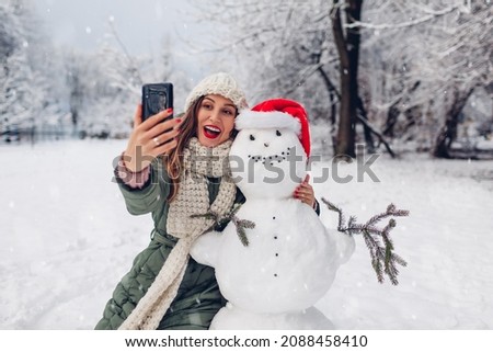 Happy woman takes selfie by snowman in Santa hat using phone outdoors in snowy winter park. Christmas festive season. Fun activities