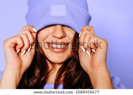 Happy young female in trendy yellow sweatshirt and knitted hat covering eyes smiling brightly against very peri background Royalty-Free Stock Photo #2088458677