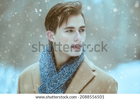 Fashion shot of a stylish handsome young man in a trendy coat and scarf posing outdoor on a snowy day. Men's beauty and fashion. Royalty-Free Stock Photo #2088556501