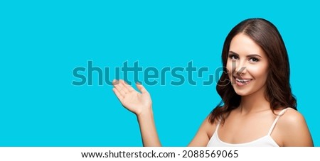 Photo of smiling beautiful woman in white casual wear holding hand with flat palm, showing, giving, advertising some product or copy space for ad text. Aqua blue green colour background. Brunette girl