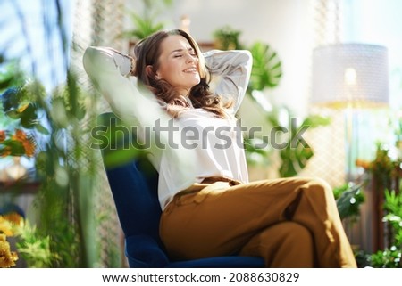 Green Home. relaxed stylish woman with long wavy hair at modern home in sunny day in green pants and grey blouse sitting in a blue armchair. Royalty-Free Stock Photo #2088630829