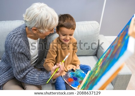 Grandson and grandma painting on canvas. Artist teaching young boy how to paint at home. Happy grandmother and a little boy painting. Senior woman with child painting on canvas. 