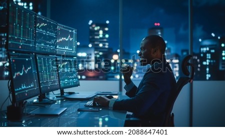 Stock Market Day Trader Working on Computer with Multi-Monitor Workstation with Real-Time Investmentment Charts. Successful African American Businessman Punches Air for Winning a Trade. Royalty-Free Stock Photo #2088647401