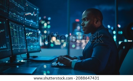 Financial Analyst Working on Computer with Multi-Monitor Workstation with Real-Time Stocks, Commodities and Exchange Market Charts. African American Trader Works in Investment Bank Late at Night. Royalty-Free Stock Photo #2088647431