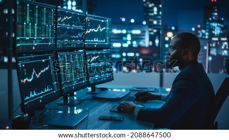 Financial Analyst Working on Computer with Multi-Monitor Workstation with Real-Time Stocks, Commodities and Exchange Market Charts. African American Trader Works in Investment Bank Late at Night. Royalty-Free Stock Photo #2088647500