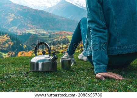 Close-up of a woman in jeans with a teapot and a cup of coffee sitting on the edge and watching the amazing beautiful mountain landscape on an autumn day.Concept of travel, outdoor activities
